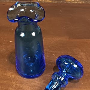 May include: A cobalt blue glass bottle with a flared top and a matching stopper. The bottle has a cylindrical shape, and the stopper has a rounded top with a stem. The glass is transparent, allowing light to pass through.