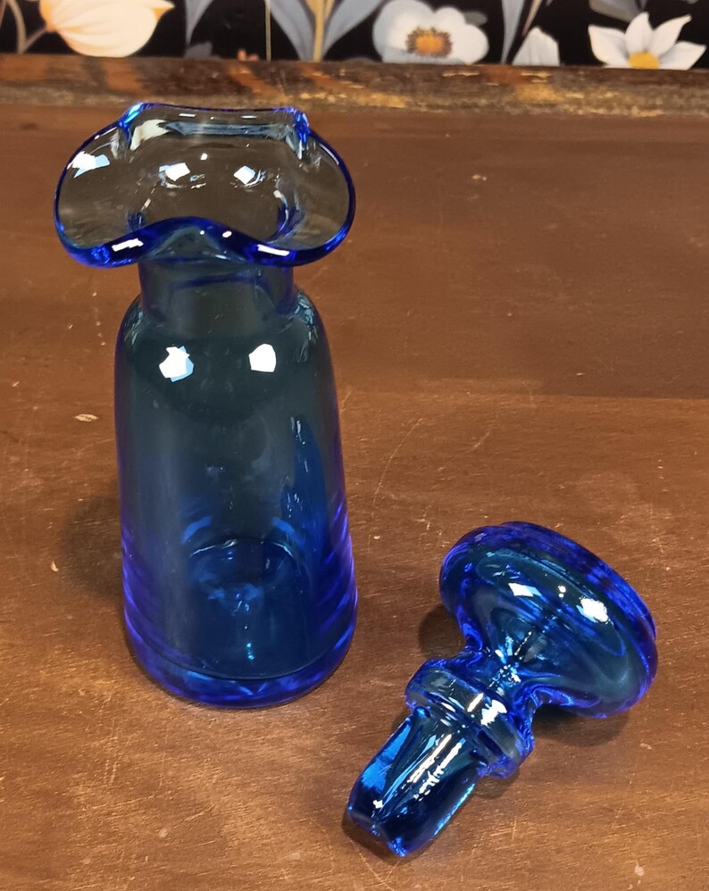 May include: A cobalt blue glass bottle with a flared top and a matching stopper. The bottle has a cylindrical shape, and the stopper has a rounded top with a stem. The glass is transparent, allowing light to pass through.