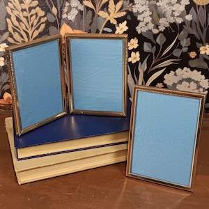 May include: Three gold-colored picture frames with light blue backing. Two frames are hinged together, and the third is separate. The frames are on top of a stack of books with a dark blue cover and cream-colored pages.
