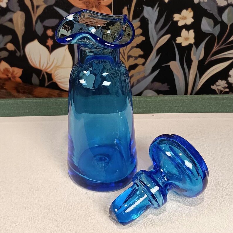 May include: A vibrant blue glass vase with a flared top and a matching stopper. The vase has a rounded body and a ruffled rim. The stopper is lying next to the vase. The background features a floral pattern.