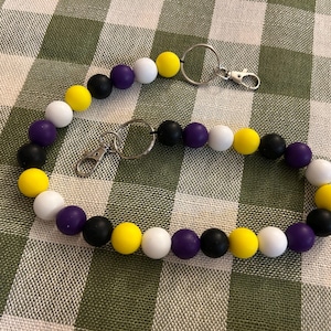 Puede incluir: Un cordón compuesto por una serie de cuentas redondas de silicona en colores alternos amarillo, blanco, morado y negro. La tira de cuentas cuenta con dos anillas de metal plateado y dos cierres de mosquetón fijados en diferentes puntos.