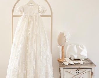 May include: A white lace christening gown with short sleeves hangs on a wooden arch. A matching bonnet, bloomers, and shoes sit on a distressed white nightstand. The items are all white, creating a soft and elegant aesthetic.