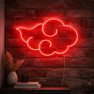Op de afbeelding: Een rode neon wolk lichtarmatuur aan een rode bakstenen muur. Het wolkenontwerp heeft een witte omtrek en een krullend ontwerp. Een vaas met gedroogde planten, boeken en een zwarte kaars staan op een tafel onder het licht.