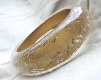 Könnte beinhalten: Ein transparentes, bernsteinfarbenes Armreif-Armband. Das Armband hat eine rissige, strukturierte Oberfläche und eine glatte, abgerundete Innenseite. Das Licht fängt die Oberfläche ein und erzeugt Glanzlichter und Schatten. Der Schmuck ist ein schlichtes, elegantes Accessoire.