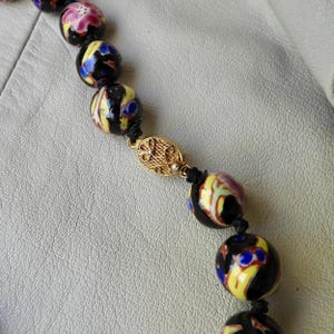May include: A vintage necklace featuring large, round beads with colorful floral and abstract designs. The beads are primarily black with accents of yellow, blue, pink, and green. A gold-toned clasp secures the necklace.