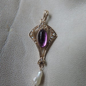 Puede incluir: Colgante dorado con una gema de amatista ovalada, rodeada de peque&ntilde;as perlas. Una perla en forma de l&aacute;grima cuelga en la parte inferior. El colgante tiene un dise&ntilde;o Art D&eacute;co ornamentado. El fondo es beige claro.