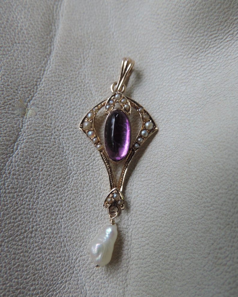 Puede incluir: Colgante dorado con una gema de amatista ovalada, rodeada de peque&ntilde;as perlas. Una perla en forma de l&aacute;grima cuelga en la parte inferior. El colgante tiene un dise&ntilde;o Art D&eacute;co ornamentado. El fondo es beige claro.