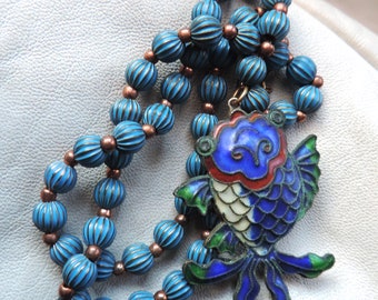 May include: A blue and white enamel fish pendant hangs from a necklace of blue and copper beads. The fish has a detailed design with green and red accents.