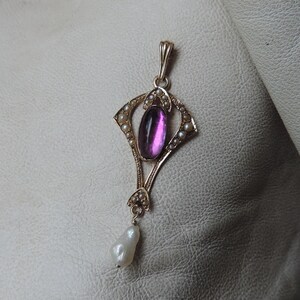 Puede incluir: Colgante de oro con una gema magenta ovalada en el centro, rodeada de peque&ntilde;as perlas. Una perla en forma de l&aacute;grima cuelga en la parte inferior. El colgante presenta un dise&ntilde;o Art Nouveau ornamentado.