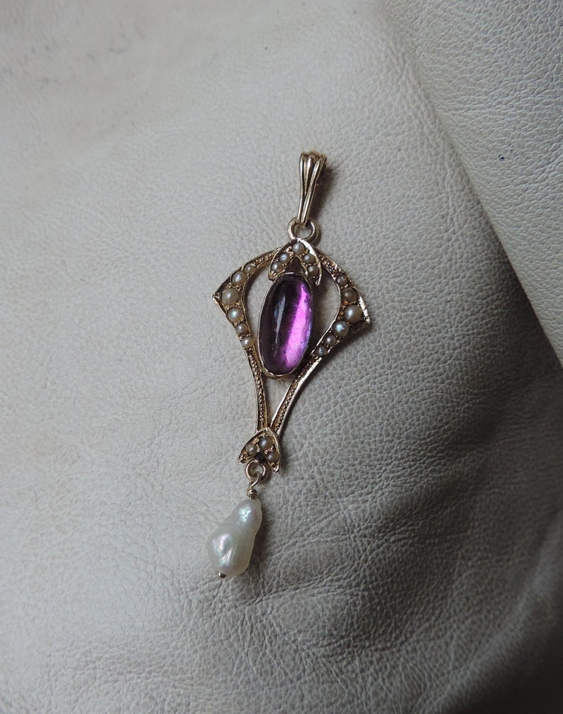 Puede incluir: Colgante de oro con una gema magenta ovalada en el centro, rodeada de peque&ntilde;as perlas. Una perla en forma de l&aacute;grima cuelga en la parte inferior. El colgante presenta un dise&ntilde;o Art Nouveau ornamentado.