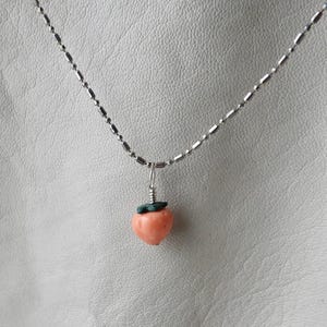 May include: A silver-toned necklace featuring a small, peach-colored strawberry pendant with green leaves. The pendant is attached to a delicate chain, creating a charming and whimsical accessory. The necklace is displayed against a neutral background.