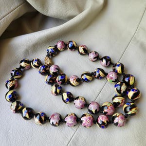 May include: A beaded necklace featuring round, hand-painted beads in black, yellow, pink, and blue floral designs. The necklace is strung on a black cord and has a gold-colored clasp. The beads are approximately 0.75 inches in diameter.