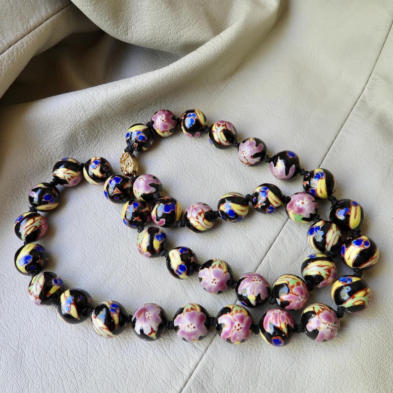 May include: A beaded necklace featuring round, hand-painted beads in black, yellow, pink, and blue floral designs. The necklace is strung on a black cord and has a gold-colored clasp. The beads are approximately 0.75 inches in diameter.