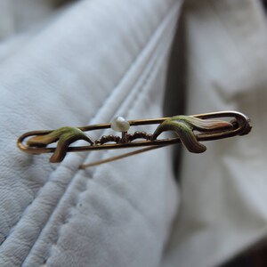 May include: A gold-toned brooch with a pearl in the center and two green enamel leaves on either side.