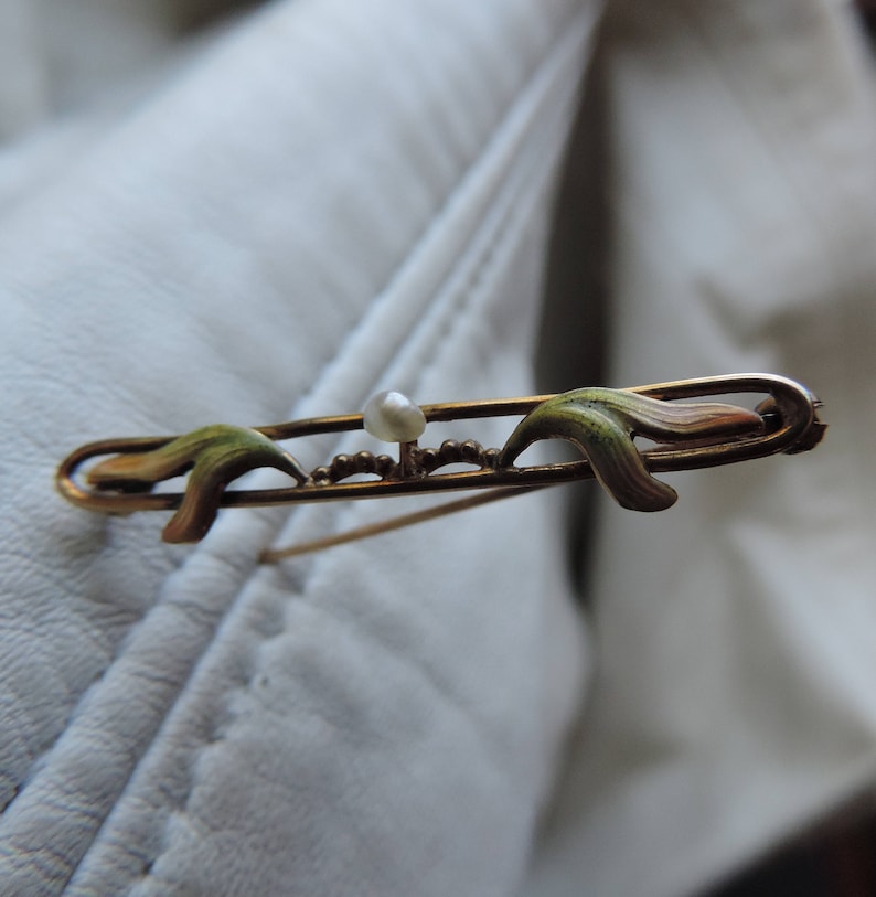 May include: A gold-toned brooch with a pearl in the center and two green enamel leaves on either side.