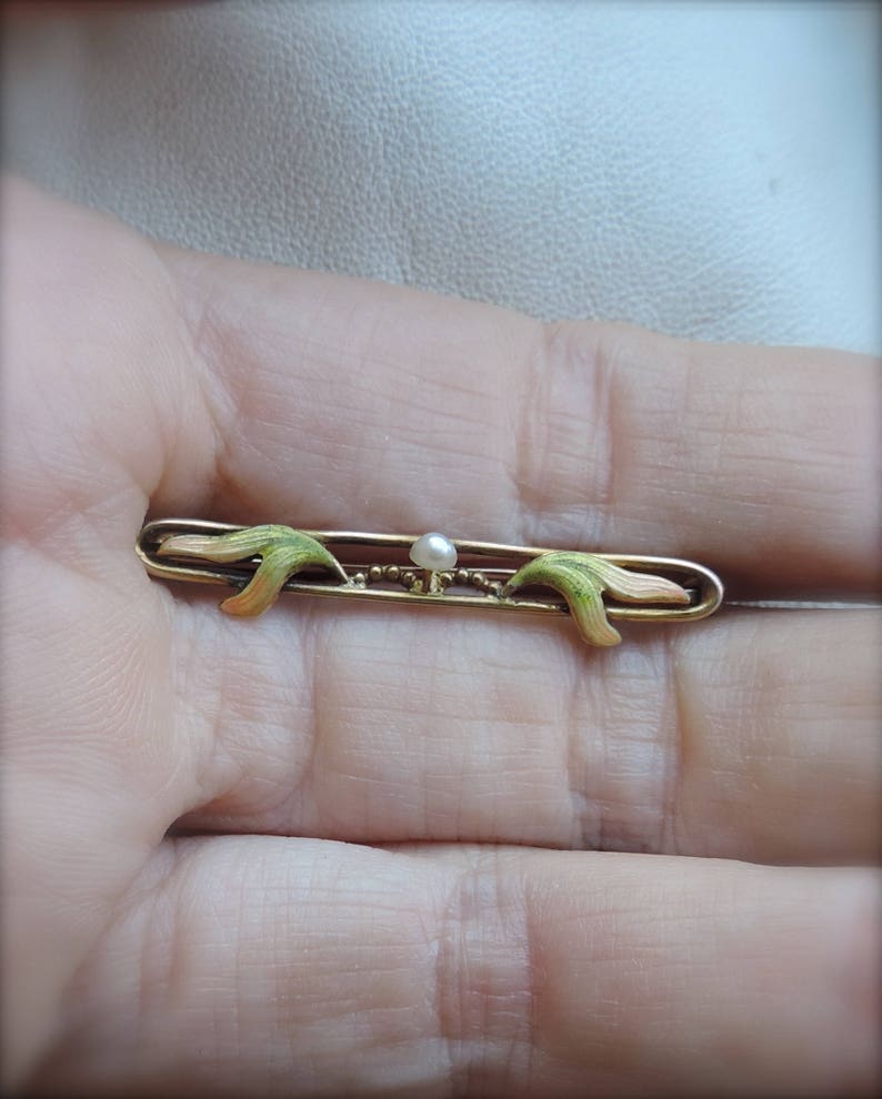 May include: A gold bar pin with a pearl in the center and green and pink enamel accents.