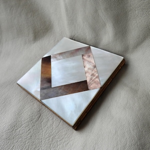 May include: Square compact with a mother-of-pearl and geometric design. The compact features a white, brown, and pink pattern with a gold-coloured metal frame. The design is a mix of natural and geometric elements.