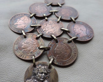 Op de afbeelding: Een kettingketting gemaakt van gekoppelde koperen munten met verschillende historische figuren. De munten zijn verbonden door kleine metalen ringen. De ketting heeft een sleutelring aan de bovenkant.