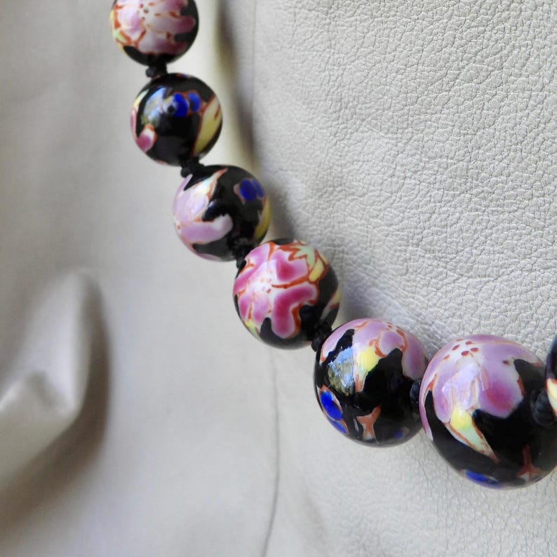 May include: A close-up of a beaded necklace featuring large, round beads. The beads are primarily black with a floral design in shades of pink, yellow, and blue. The necklace is strung on a black cord.