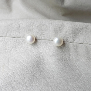 May include: Two round, white pearl earrings resting on a textured, off-white surface. The pearls are smooth and luminous, reflecting light. The background is a close-up of a leather-like material.