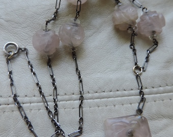 May include: A silver chain necklace with a pendant and several carved rose quartz beads. The pendant is a square shape with a carved rose design.