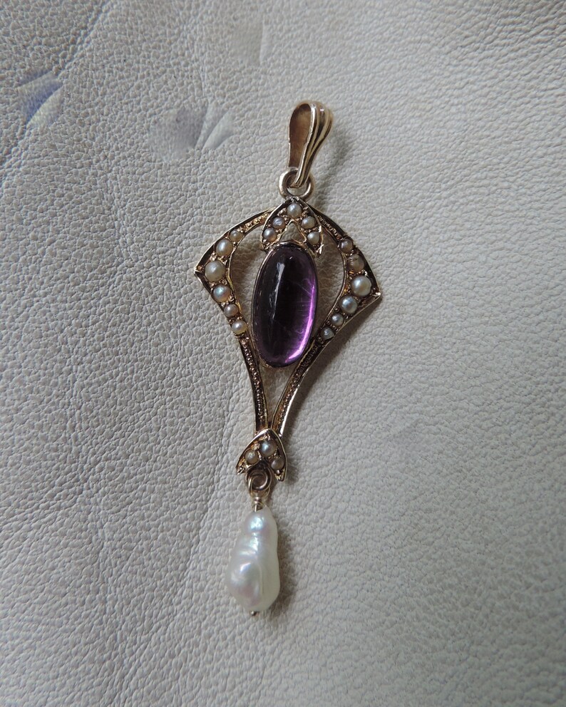 Puede incluir: Colgante de oro con una piedra amatista ovalada, rodeada de peque&ntilde;as perlas. Una perla en forma de l&aacute;grima cuelga de la parte inferior. El colgante tiene un dise&ntilde;o Art Nouveau ornamentado.