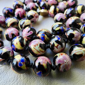 May include: A close-up of a necklace featuring round, colorful beads. The beads have a black base with swirls of yellow, pink, and blue. The necklace is strung on a black cord and has a gold-colored clasp.