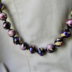 May include: A beaded necklace featuring large, round beads. The beads are primarily black with floral and abstract designs in pink, yellow, and blue. The necklace is strung on a black cord.