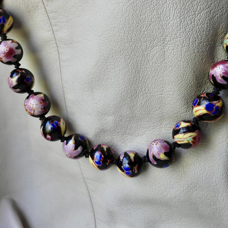 May include: A beaded necklace featuring large, round beads. The beads are primarily black with floral and abstract designs in pink, yellow, and blue. The necklace is strung on a black cord.