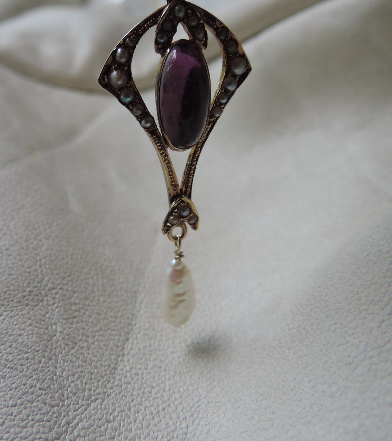 Puede incluir: Colgante dorado con una gema ovalada de color p&uacute;rpura oscuro en el centro. El colgante est&aacute; adornado con peque&ntilde;as piedras redondas y claras, y una perla en forma de l&aacute;grima. El dise&ntilde;o es intrincado y elegante.
