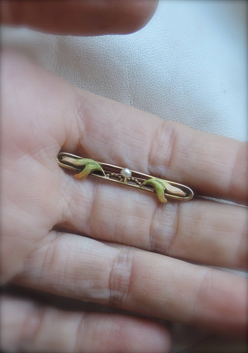 May include: A gold-toned brooch with a delicate design featuring two green and pink leaves on either side of a single pearl.