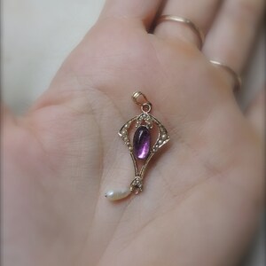Puede incluir: Colgante de oro con una piedra preciosa ovalada p&uacute;rpura y una peque&ntilde;a gota de perla blanca. El colgante est&aacute; rodeado de peque&ntilde;as perlas y tiene un dise&ntilde;o Art Nouveau ornamentado.
