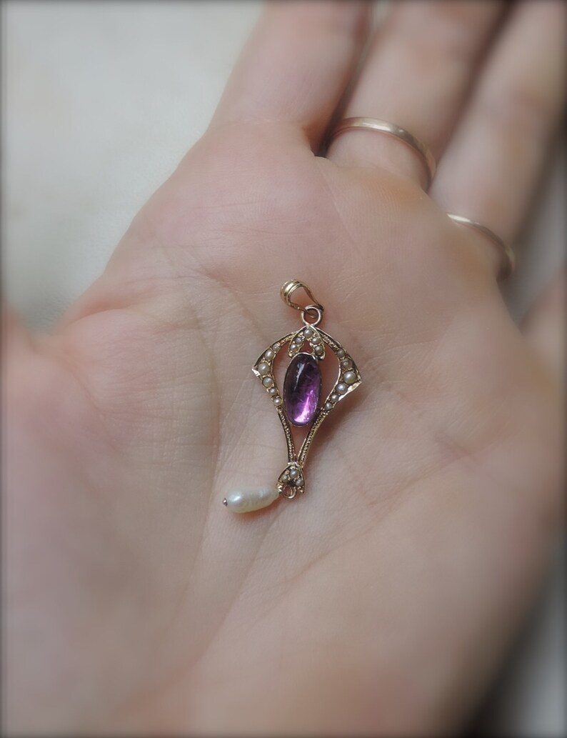 Puede incluir: Colgante de oro con una piedra preciosa ovalada p&uacute;rpura y una peque&ntilde;a gota de perla blanca. El colgante est&aacute; rodeado de peque&ntilde;as perlas y tiene un dise&ntilde;o Art Nouveau ornamentado.