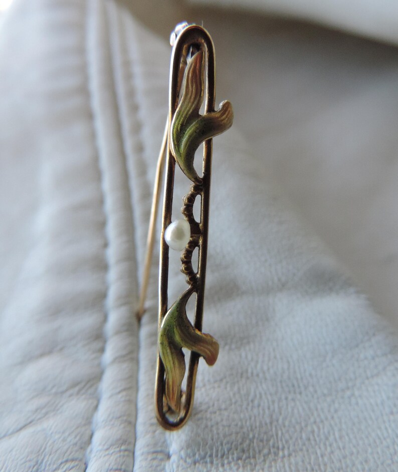 May include: A gold-toned brooch with a delicate design featuring two stylized leaves and a single pearl. The leaves are enameled in shades of green and gold.