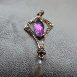 Puede incluir: Colgante de oro con una gran gema rosa ovalada en el centro, rodeada de peque&ntilde;as perlas. Una perla en forma de l&aacute;grima cuelga en la parte inferior. El colgante tiene un dise&ntilde;o Art Nouveau ornamentado.