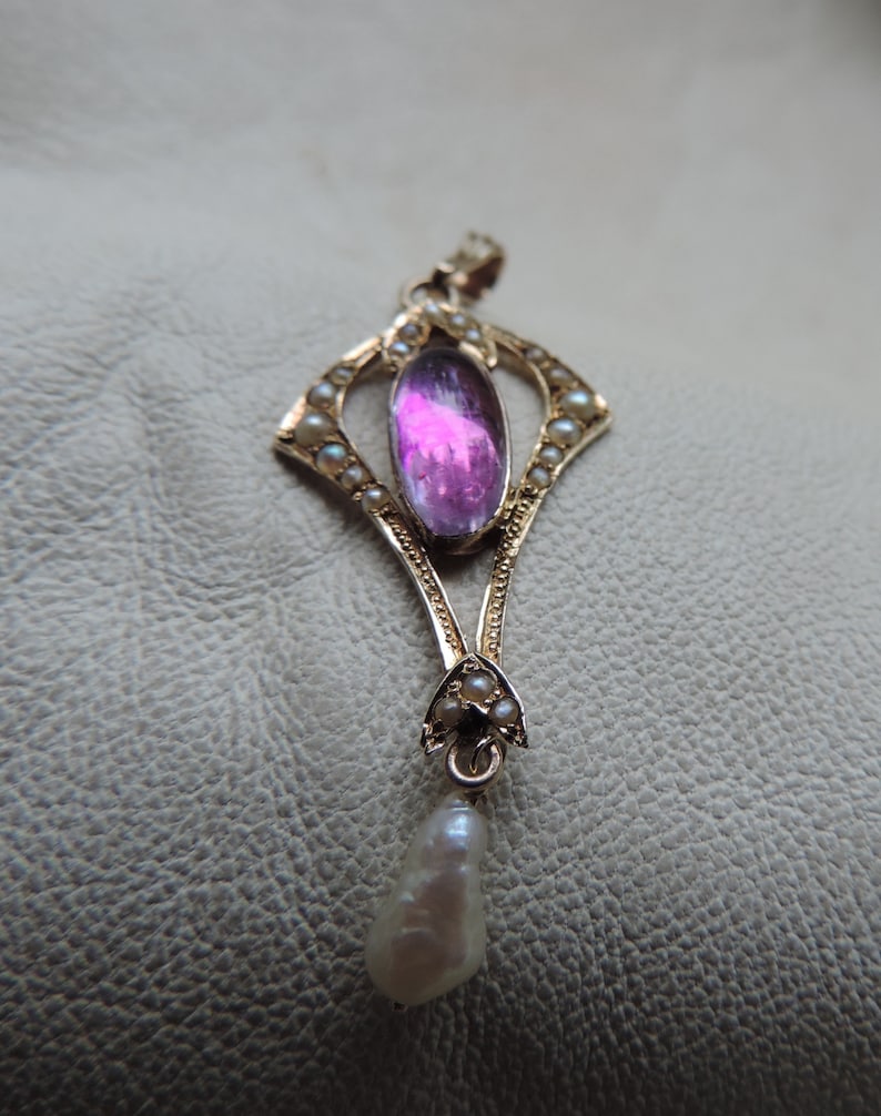 Puede incluir: Colgante de oro con una gran gema rosa ovalada en el centro, rodeada de peque&ntilde;as perlas. Una perla en forma de l&aacute;grima cuelga en la parte inferior. El colgante tiene un dise&ntilde;o Art Nouveau ornamentado.