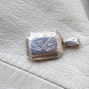 May include: A rectangular silver pendant with intricate floral engraving. The pendant has a small loop for a chain and is set against a textured, off-white surface. The design features detailed patterns and a polished finish.