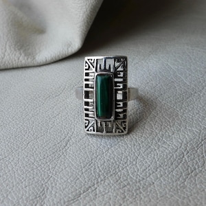 Könnte beinhalten: Ein Silberring mit einem rechteckigen Malachitstein, der in einem dekorativen Rahmen gefasst ist. Der Ring hat ein durchbrochenes Design um den grünen Stein. Der Ring wird auf einer strukturierten Oberfläche präsentiert.