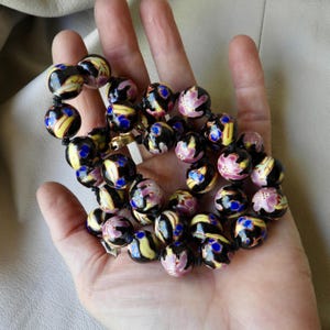 May include: A necklace of black, round beads with floral and abstract designs in yellow, pink, and blue. The beads are strung on a black cord and held in an open hand.