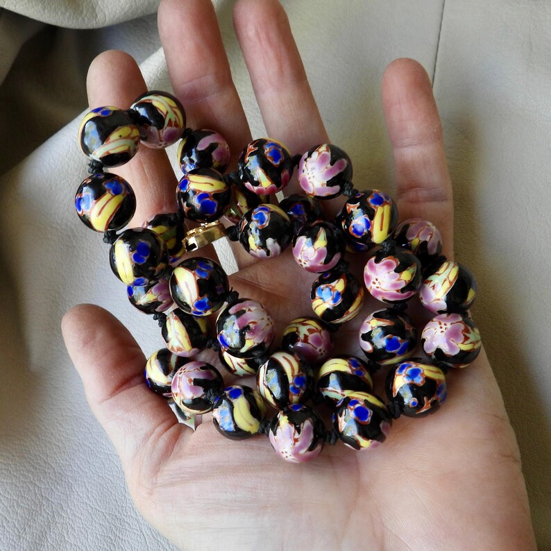 May include: A necklace of black, round beads with floral and abstract designs in yellow, pink, and blue. The beads are strung on a black cord and held in an open hand.