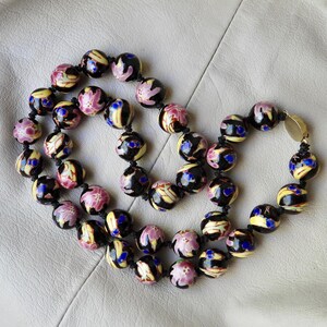 May include: A beaded necklace featuring round, black beads with floral and abstract designs in pink, yellow, and blue. The necklace is strung on a black cord and has a gold-colored clasp.