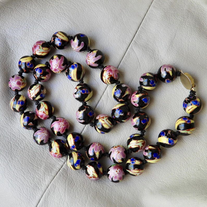 May include: A beaded necklace featuring round, black beads with floral and abstract designs in pink, yellow, and blue. The necklace is strung on a black cord and has a gold-colored clasp.