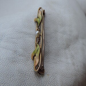 May include: A gold-toned brooch with a delicate design of green and pink leaves and a single pearl in the center.
