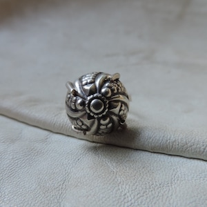 May include: A silver ring with a detailed, ornate design. The ring features a round, textured centre with a raised, circular pattern.