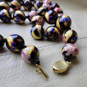 May include: A beaded necklace featuring large, round beads in a variety of colors. The beads have a black base with yellow, blue, and pink floral patterns. The necklace is strung on a black cord and has a gold-colored clasp.
