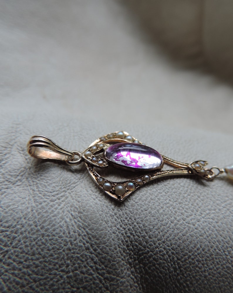 Puede incluir: Colgante dorado con una gran gema rosa ovalada en el centro. El colgante est&aacute; adornado con peque&ntilde;as perlas y detalles de filigrana intrincados. El dise&ntilde;o es ornamentado y de inspiraci&oacute;n vintage, con una delicada cadena.