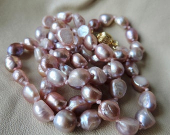 May include: A long strand of lavender-colored freshwater pearls, knotted and strung on a thin thread. The pearls are irregular in shape and have a subtle iridescent sheen.