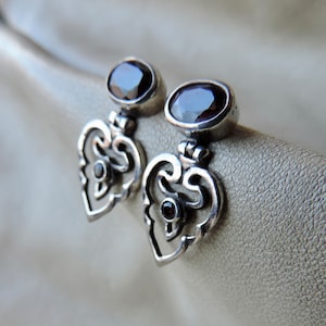 May include: A pair of silver earrings with a heart-shaped design and dark purple gemstones. The earrings have a delicate, intricate design.