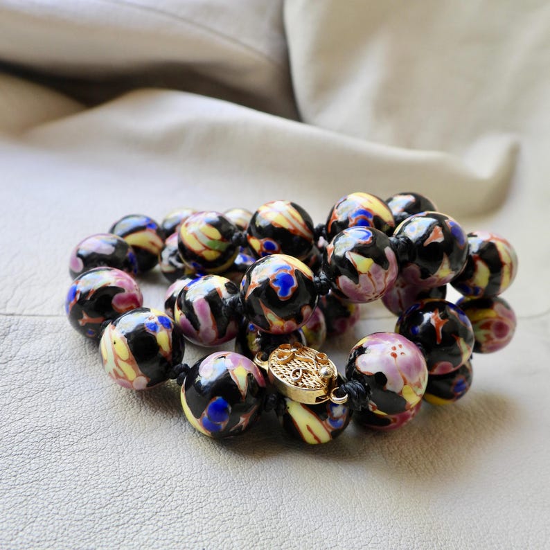 May include: A beaded necklace featuring large, round beads with a black base and colorful swirls of yellow, pink, and blue. The necklace is fastened with a gold-colored clasp. The beads are approximately 1 inch in diameter.