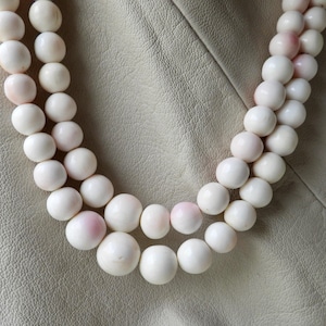 Peut inclure: Collier à deux rangs de perles blanches, légèrement rosées. Les perles rondes sont de taille uniforme et rapprochées, créant un aspect plein et élégant. Le collier est présenté sur un fond beige clair.